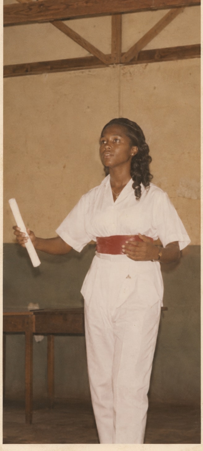 Oby as a schoolgirl standing in a hall in white with a red sash, a rolled scroll in her raised hand, mid-speech