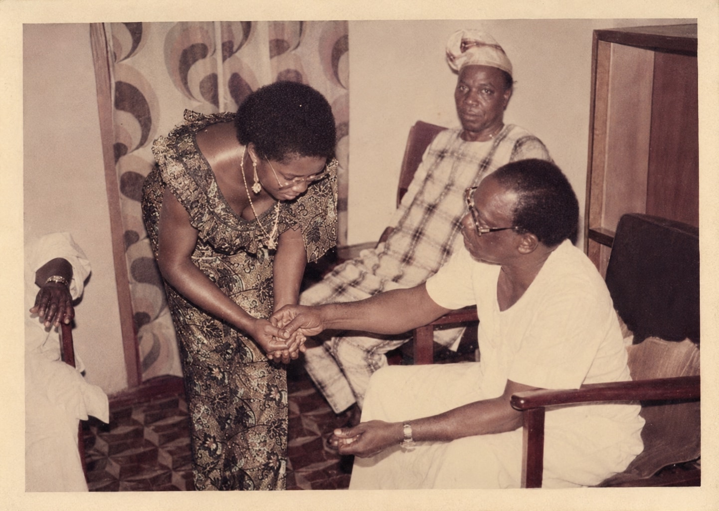 Oby's father seated indoors in white, holding hands with her mother, who is bending towards him