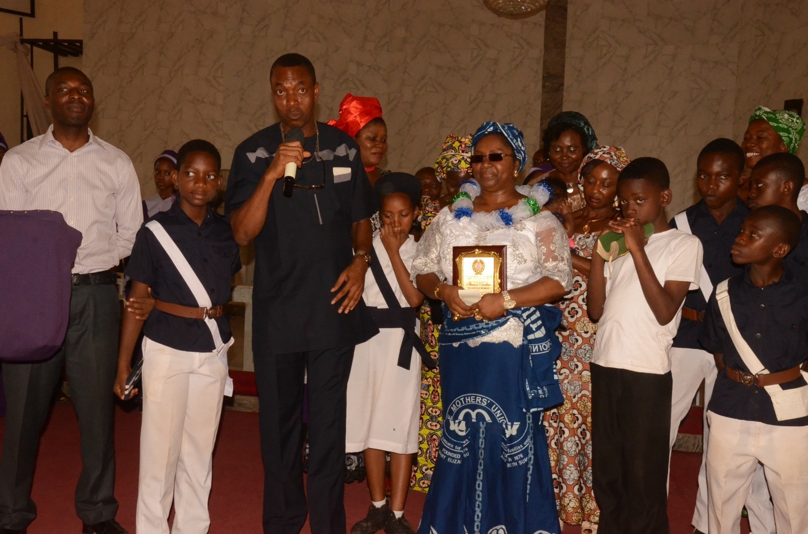 A hall of children in uniform and sashes at a presentation, Dr. Oby Nwokedi among the adults