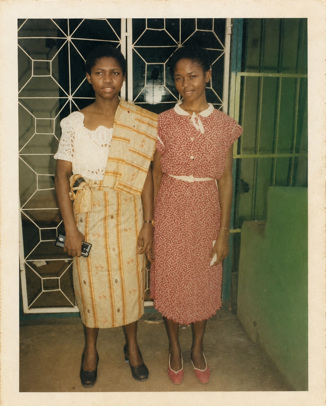 Oby and her childhood friend Onyechi standing together in front of a green door, both in patterned dress