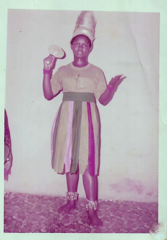 Oby as a girl in her school dance group costume, a tall conical headdress and rattles at her ankles