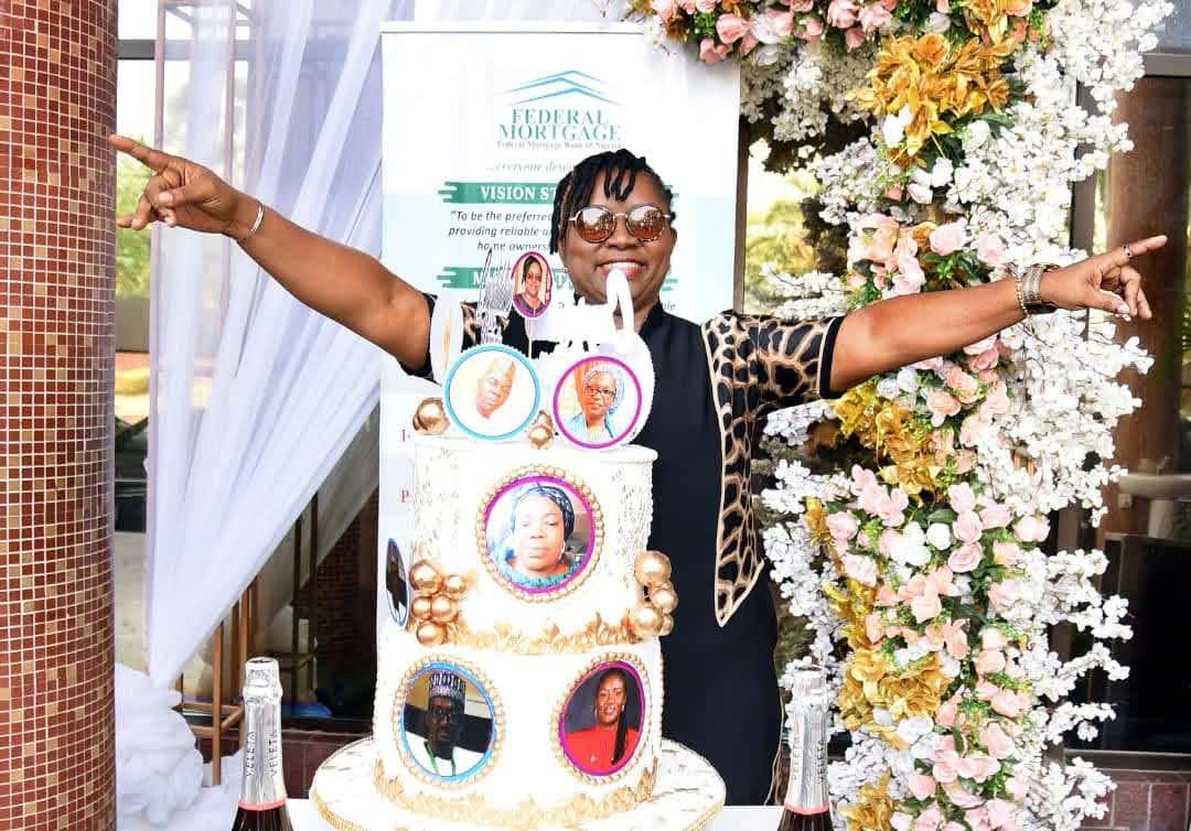 Dr. Oby Nwokedi with both arms flung wide behind a two-tier cake printed with portraits, before a Federal Mortgage Bank banner
