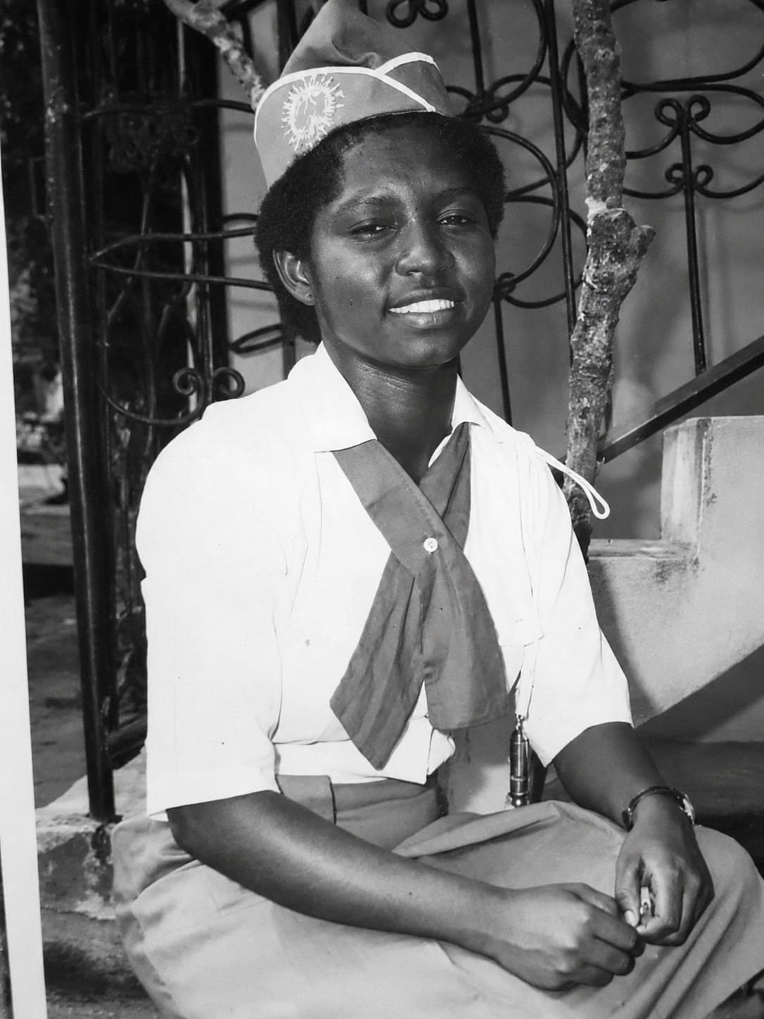 Oby Attoh as a schoolgirl in girl guide uniform, seated on a step before an iron gate