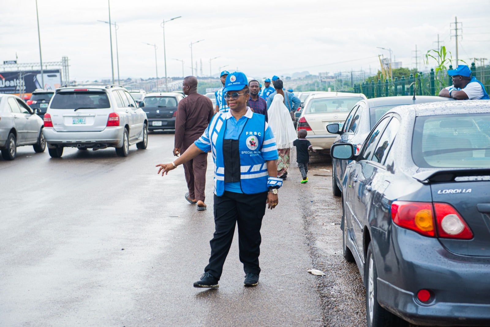 Dr. Oby Nwokedi in an FRSC Special Marshal vest and cap, one hand raised, directing traffic between queued cars on a wet road