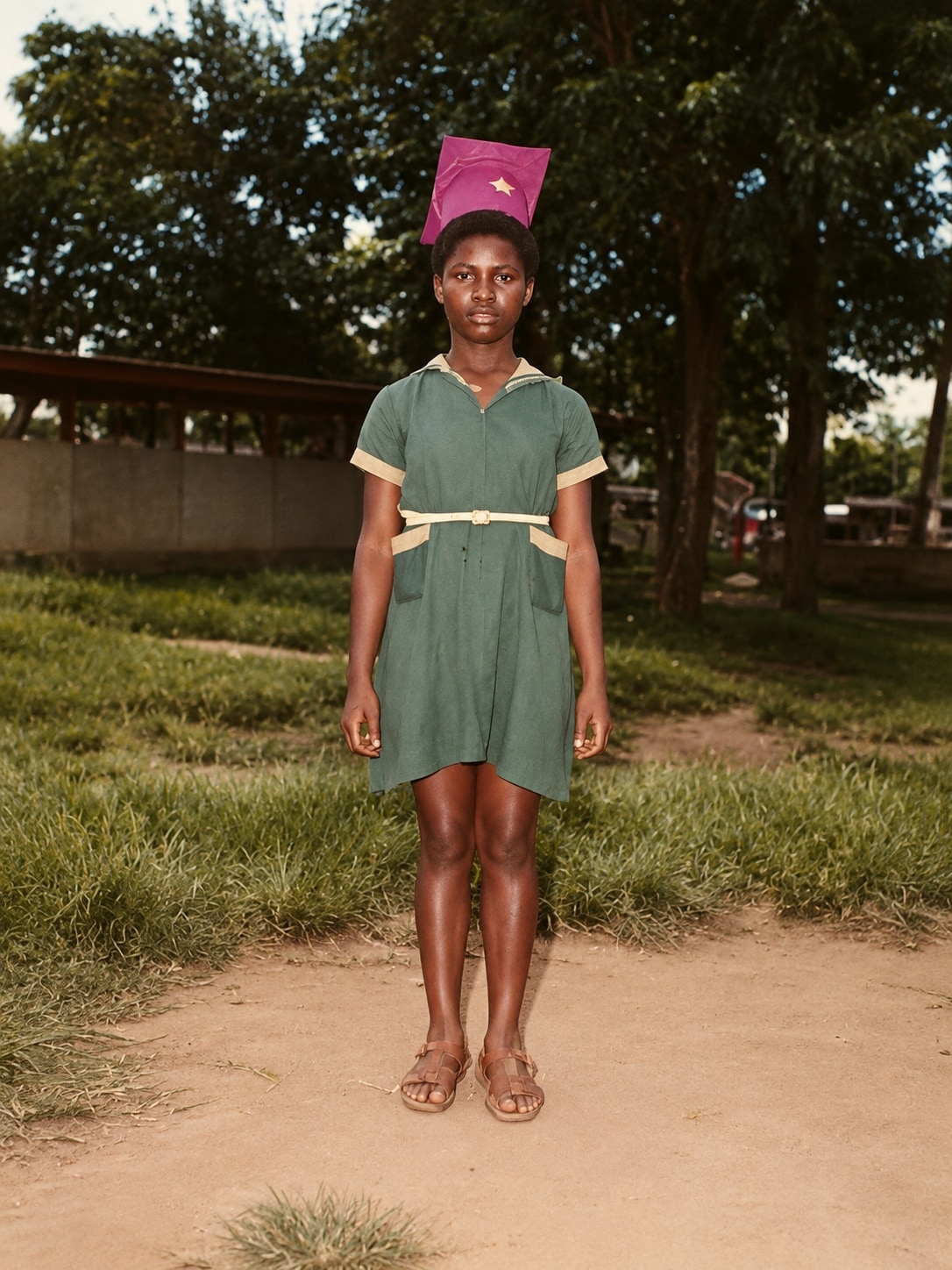 Oby as a schoolgirl in a green school dress and pink cap, standing on a path at a school event