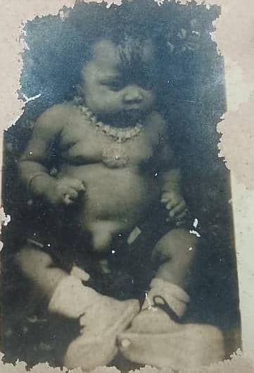 A worn black and white photograph of Oby as a baby, seated, wearing a beaded necklace and bootees